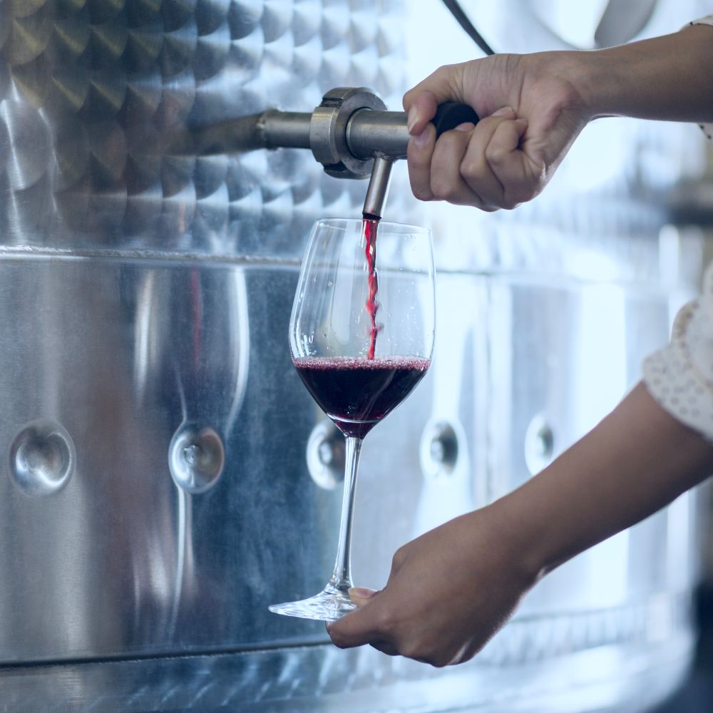 photo of a woman filling a wine glass with red wine from am industrial steel barrel