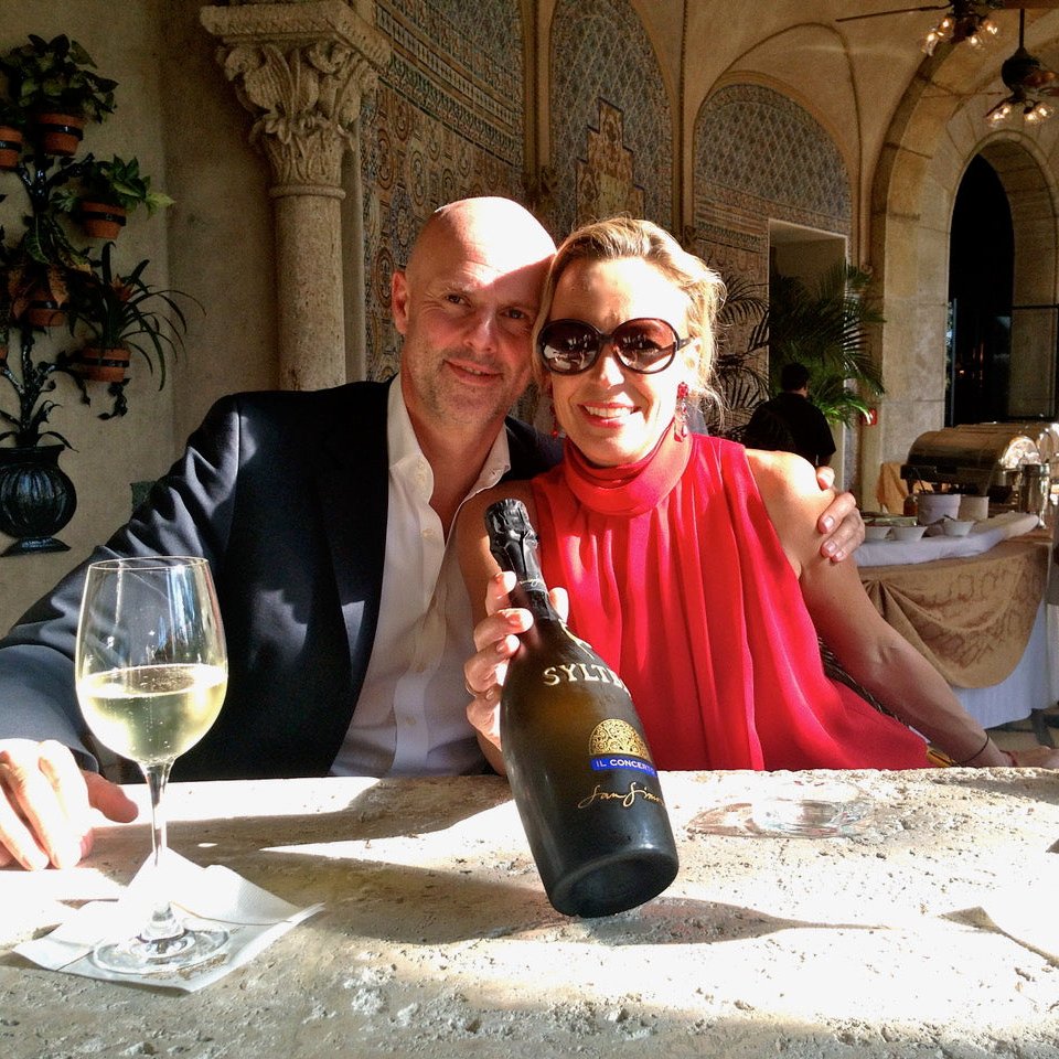 photo of SYLTBAR owners Regina and Claus smiling and sitting at a table at Mar-a-Lago in Palm Beach holding a bottle of Mr SYLTBAR low-sugar prosecco